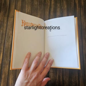 K&ouml;nnte beinhalten: Ein offenes Buch mit orangefarbenen Kanten und einem Holzuntergrund. Auf der linken Seite steht das Wort "Halloween" in orangefarbener Schrift. Die rechte Seite ist leer. Der Text "starlightcreations" ist ebenfalls sichtbar. Eine Hand ist teilweise sichtbar.