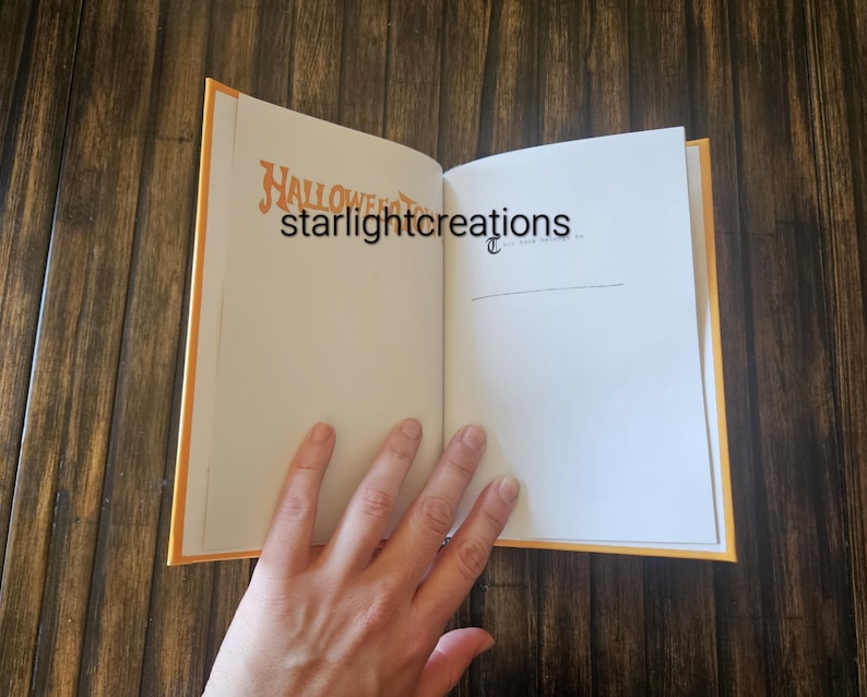 K&ouml;nnte beinhalten: Ein offenes Buch mit orangefarbenen Kanten und einem Holzuntergrund. Auf der linken Seite steht das Wort "Halloween" in orangefarbener Schrift. Die rechte Seite ist leer. Der Text "starlightcreations" ist ebenfalls sichtbar. Eine Hand ist teilweise sichtbar.