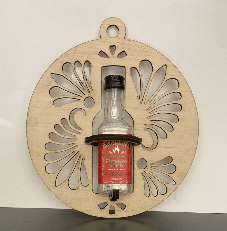 May include: A wooden ornament shaped like a circle with a decorative design. A small bottle of Jack Daniel's Tennessee Fire whiskey is displayed in the center of the ornament.
