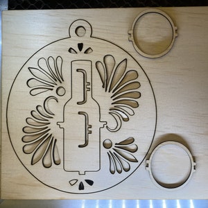 May include: A laser-cut wooden ornament in the shape of a circle with a detailed design of a bottle and stylized floral patterns. The ornament has a hole at the top for hanging.