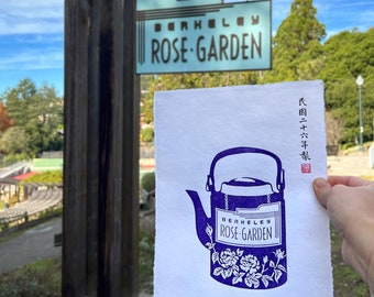 May include: A purple and white illustration of a teapot with the text "Berkeley Rose Garden" on the side. The illustration is on a white piece of paper with the text "Berkeley Rose Garden Since 1913" at the bottom. The image is in front of a wooden sign that reads "Berkeley Rose Garden".