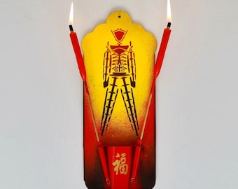 May include: A decorative wall hanging featuring a stylized figure with a yellow and red color scheme. Two red candles are lit, flanking the central design. The base includes a red container with gold Chinese characters.
