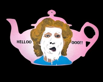 May include: A pink teapot-shaped art piece with a portrait of a person with a white face mask and brown curly hair. The text "HELLOO OOO!!" is printed on the teapot. The background is black.