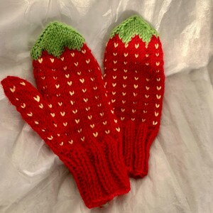 May include: A pair of hand-knitted mittens featuring a red strawberry pattern with small yellow V-shaped stitches. The top of each mitten has a green knitted cuff resembling strawberry leaves, and the wrist area features a ribbed red knit texture.
