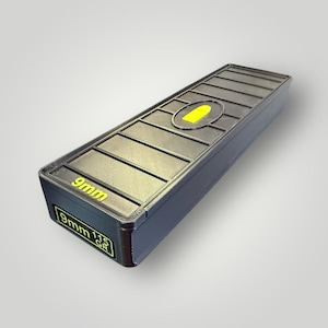 May include: A dark grey rectangular container with a textured lid. The lid features a yellow bullet-shaped design and the text "9mm". The side of the container also displays "9mm 115 GR" in yellow. The container is designed to hold ammunition.