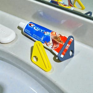 Puede incluir: Un tubo de pasta de dientes Crest azul y blanco con un exprimidor de tubos de plástico gris y un exprimidor de plástico amarillo. El tubo de pasta de dientes descansa sobre una superficie blanca. El exprimidor de tubos tiene el texto "NA PRES JONES".