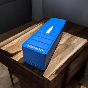 Könnte beinhalten: Ein blauer rechteckiger Behälter mit dem Text "5.56 NATO" auf der Vorder- und Seite. Die Oberseite hat ein erhabenes Design mit einer weißen Kugelform. Der Behälter steht auf einer Holzoberfläche.