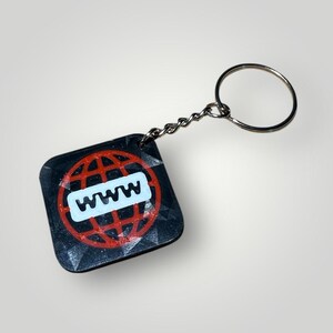 Op de afbeelding: Zwarte vierkante sleutelhanger met een rood wereldbolontwerp en de witte letters "WWW". De sleutelhanger heeft een zilveren ketting en ring. Het ontwerp staat op een getextureerd oppervlak.