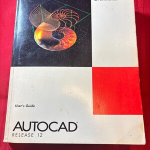 May include: A vintage AutoCAD Release 12 User's Guide book. The cover features a geometric design in orange, green, and white on a black background. The upper right corner has the Autodesk logo and a red square.