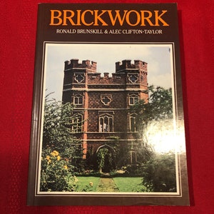 May include: A hardcover book titled "BRICKWORK" by Ronald Brunskill & Alec Clifton-Taylor. The cover features a color image of a brick building with a castle-like design, surrounded by greenery. The book is set against a red background.