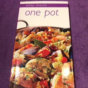 Könnte beinhalten: Ein Kochbuch mit dem Titel "easy meals one pot" zeigt ein lebendiges Bild eines Eintopfgerichts mit Meeresfrüchten, Hühnchen und Gemüse. Das Cover zeigt eine Nahaufnahme des gekochten Gerichts mit Reis, Garnelen, Muscheln und roten Paprika.