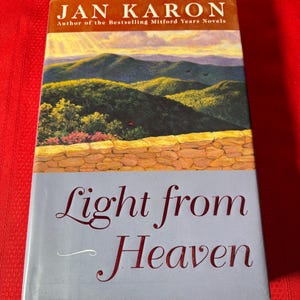 May include: A book titled "Light from Heaven" by Jan Karon, author of the bestselling Mitford Years novels. The cover features a painting of a mountain range with a stone wall in the foreground.