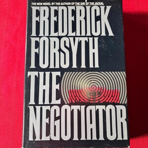 Op de afbeelding: Boekomslag van "The Negotiator" van Frederick Forsyth. De zwarte cover heeft witte letters met de titel en de naam van de auteur. Een spiraalvormig ontwerp in rood en wit staat centraal.