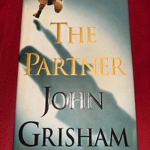 May include: A hardcover book titled "The Partner" by John Grisham. The cover features a shadowy figure and the title in gold lettering. The background is a gradient of blue and green.
