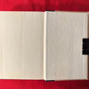 May include: A blank, white book with a brown spine is open to a blank page. The book is lying flat on a red surface.
