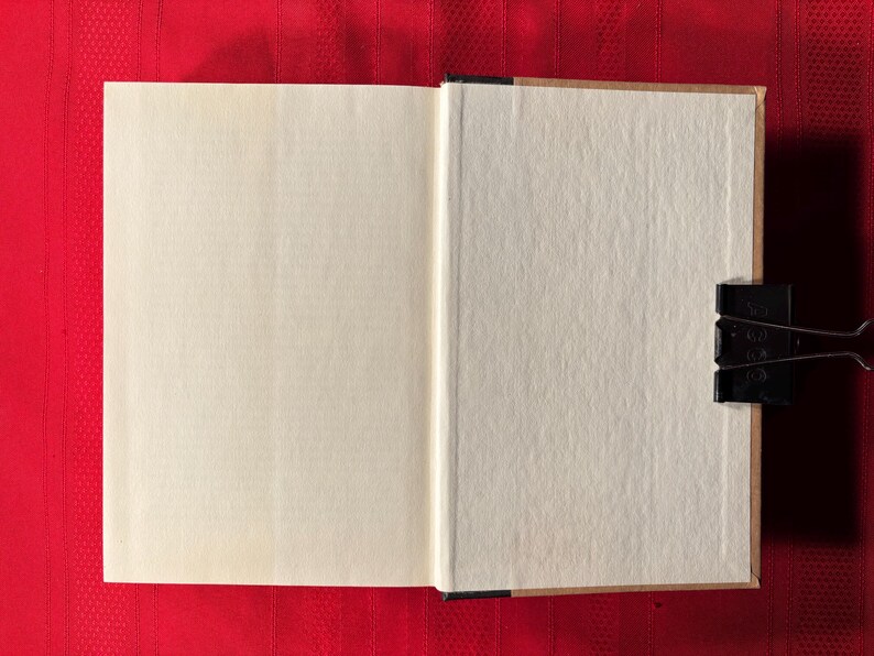 May include: A blank, white book with a brown spine is open to a blank page. The book is lying flat on a red surface.
