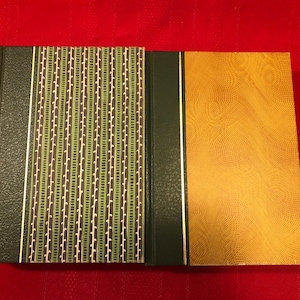 Könnte beinhalten: Zwei gebundene Bücher mit grünen und goldenen Einbänden. Das Buch links hat ein Muster mit grünen, weißen und schwarzen Streifen. Das Buch rechts hat einen einfarbigen goldenen Einband mit einem dezenten strukturierten Muster.