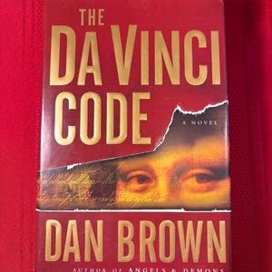 Op de afbeelding: Een rood hardcover boek, "The Da Vinci Code" van Dan Brown, met gouden letters. De cover toont een afbeelding van ogen en een gescheurd papiereffect, wat een gelaagde uitstraling creëert.