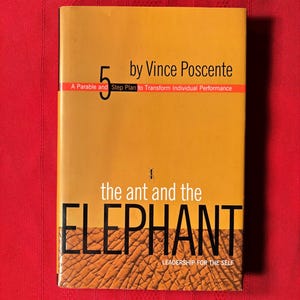 Op de afbeelding: Een geel boek getiteld "De mier en de olifant: Leiderschap voor jezelf" door Vince Poscente. De omslag van het boek toont een kleine mier en een close-up van de huid van een olifant.