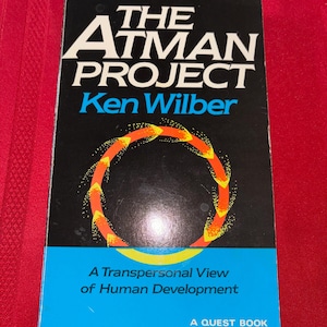 Puede incluir: Un libro de bolsillo titulado "The Atman Project" de Ken Wilber. La portada presenta texto blanco sobre un fondo negro, con un diseño circular en naranja y amarillo. El subtítulo del libro es "A Transpersonal View of Human Development."