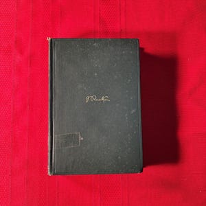 Könnte beinhalten: Ein dunkelgrünes Buch mit goldener Schrift, das auf einer roten Oberfläche liegt. Der Einband zeigt die Unterschrift des Autors in goldener Schrift. Das Buch ist leicht abgenutzt, was auf Alter und Nutzung hindeutet. Die Gesamtästhetik ist klassisch und vintage.