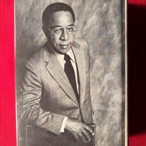 May include: A black and white photo of a man wearing a gray suit jacket, a white shirt, and a dark tie. He is wearing glasses and has a serious expression on his face. He is leaning against a dark surface.