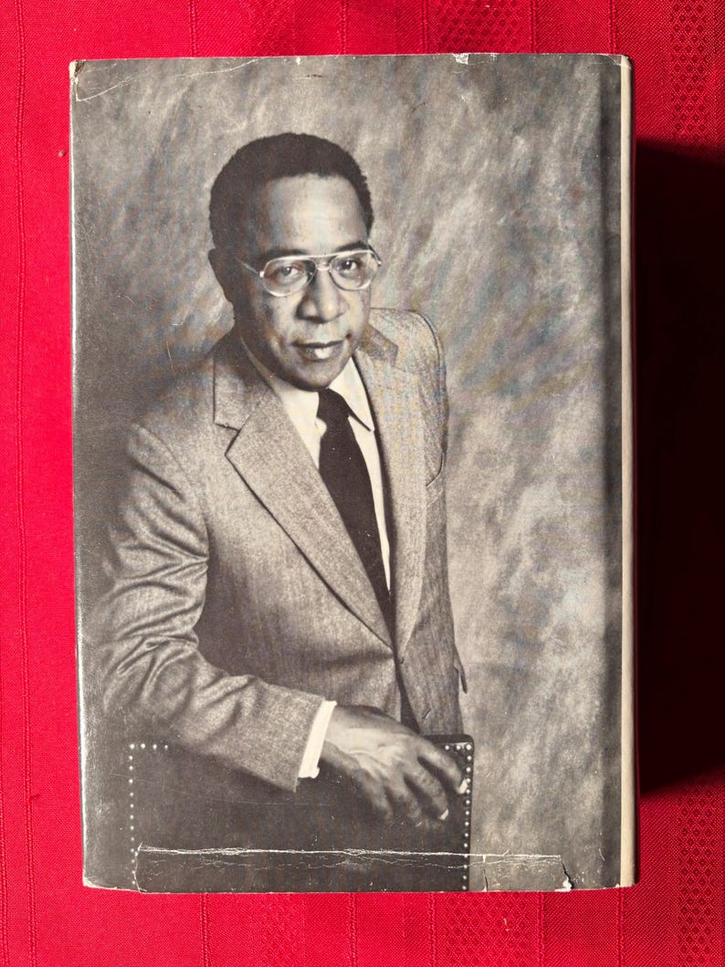 May include: A black and white photo of a man wearing a gray suit jacket, a white shirt, and a dark tie. He is wearing glasses and has a serious expression on his face. He is leaning against a dark surface.