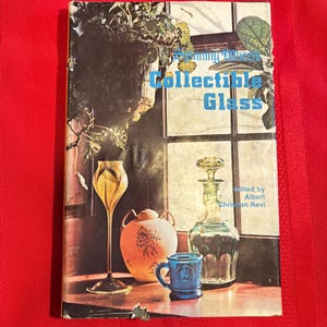 May include: A vintage book cover titled "Collecting Antique Glass" with various glass objects displayed on a table. The cover features a window with plants and a collection of glassware, including a goblet, a decanter, and a blue mug.