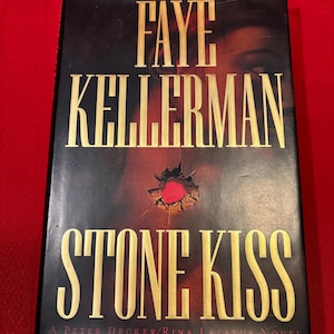 Może przedstawiać: Czarna i złota książka w miękkiej oprawie z tytułem "Stone Kiss" autorstwa Faye Kellerman. Książka jest powieścią Petera Deckera/Riny Lazarus.