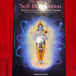 May include: A hardcover book titled The Science of Self-Realization by His Divine Grace A.C. Bhaktivedanta Swami Prabhupada. The black cover features a central illustration of a four-armed deity standing on a lotus, surrounded by blue atomic orbits.