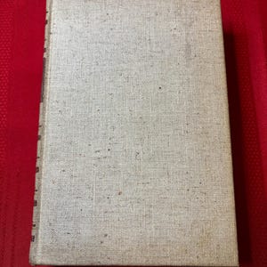 May include: A vintage hardcover book with a beige linen cover. The book has a textured surface and a slightly worn appearance.