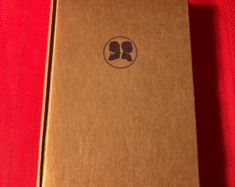 Könnte beinhalten: Ein geschlossenes Hardcover-Buch mit strukturiertem, goldfarbenem Einband. Ein kreisförmiges Emblem mit zwei Silhouetten ist mittig auf der Vorderseite. Das Buch liegt vor rotem Hintergrund.