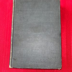 May include: A closed, hardcover book with a dark gray cover. The cover has a textured appearance and shows signs of wear along the edges. The book is resting on a red surface.