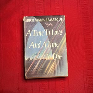 Op de afbeelding: Een vintage boek getiteld "A Time To Love And A Time To Die" van Erich Maria Remarque. De boekomslag toont een gestileerde illustratie met een boom, gebouwen en een regenboog tegen een blauw-grijze achtergrond. Het boek ligt op een rode ondergrond.