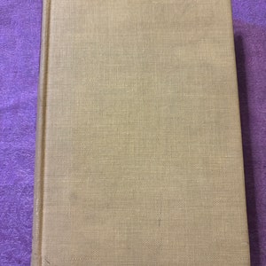 May include: A hardcover book with a beige linen cover. The book is rectangular and has a textured surface. The spine is visible on the left side of the book.