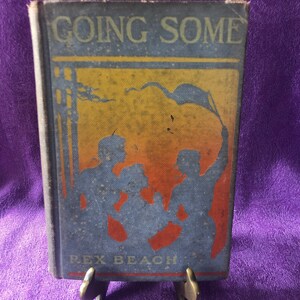 Könnte beinhalten: Ein antikes Buch mit dem Titel "Going Some" von Rex Beach. Das Cover zeigt eine blau-orangefarbene Illustration von Silhouetten und einer wehenden Flagge. Das Buch wird auf einem kleinen, dekorativen Ständer vor einem lila Hintergrund präsentiert.