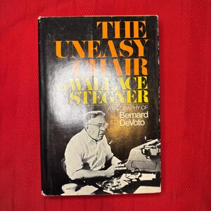 Peut inclure: Un livre vintage intitulé "The Uneasy Chair" par Wallace Stegner, avec une biographie de Bernard DeVoto. La couverture est noire avec du texte orange et blanc. L'image présente une photo en noir et blanc d'un homme devant une machine à écrire.