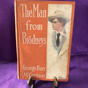 Pode incluir: Um livro antigo de capa dura intitulado "The Man from Brodneys" de George Barr McCutcheon. A capa apresenta um homem com um chapéu branco e um terno, olhando para o lado.