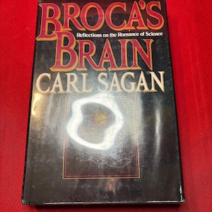 May include: A black hardback book with the title "Broca's Brain: Reflections on the Romance of Science" by Carl Sagan. The book cover features a black and white image of a nebula.