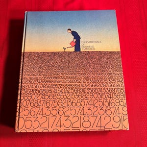 May include: A book titled "Fundamentals of Business Statistics" with a cartoon illustration of a person watering a small plant. The cover is orange with black text and numbers.