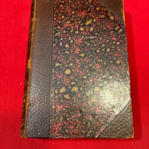 May include: A vintage, brown leather-bound book with a red and gold marbled cover. The book has a spine with raised lettering and a decorative design on the front cover.