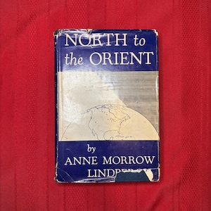 May include: A vintage book titled "NORTH to the ORIENT" by Anne Morrow Lindbergh. The book has a blue cover with white text and a world map illustration. The book is slightly worn, with visible signs of age.
