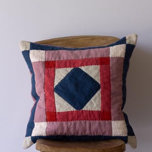 May include: Square patchwork pillow with a central blue diamond design. The pillow features a patchwork design with red, pink, white, and blue squares. The pillow is placed on a wooden chair.