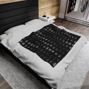 May include: A black throw blanket with a white moon phase pattern. The blanket is folded and placed on a white bed.
