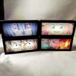 May include: Four rectangular light boxes with black frames, each displaying a different anime character illustration. The images feature a blue-haired character, a pink-haired character, a wide-eyed character, and a character with fire effects.