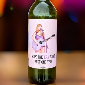 May include: Green glass wine bottle with a label featuring an illustration of a singer with a guitar. The label includes the text "I HOPE THIS ERA IS THE BEST ONE YET!"