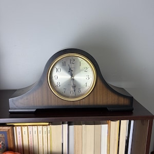 May include: A mantel clock with a dark wood grain finish and a curved top. The clock face is circular with a gold-toned rim, a silver background, black Arabic numerals, and ornate black hands. The word Linden is printed on the clock face.