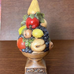 Puede incluir: Una escultura decorativa de mesa con una disposición piramidal de frutas variadas que incluye una pera amarilla, una manzana roja, un limón amarillo, una naranja y uvas de color morado oscuro, sobre una base de pedestal marrón con detalles.