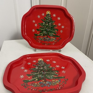 May include: Two red metal trays with scalloped edges, each featuring a central illustration of a decorated Christmas tree surrounded by gifts and white snowflakes. The trays have a glossy finish and a raised rim, displaying a festive holiday scene.