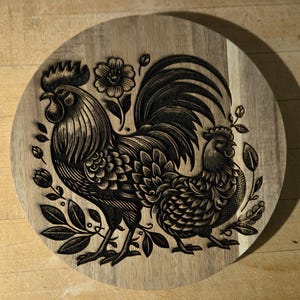 Könnte beinhalten: Ein rundes Holzschneidebrett mit einem detaillierten gravierten Design eines Hahns und einer Henne, umgeben von Blumen- und Blättermotiven. Das Brett ist aus hellem Holz gefertigt.